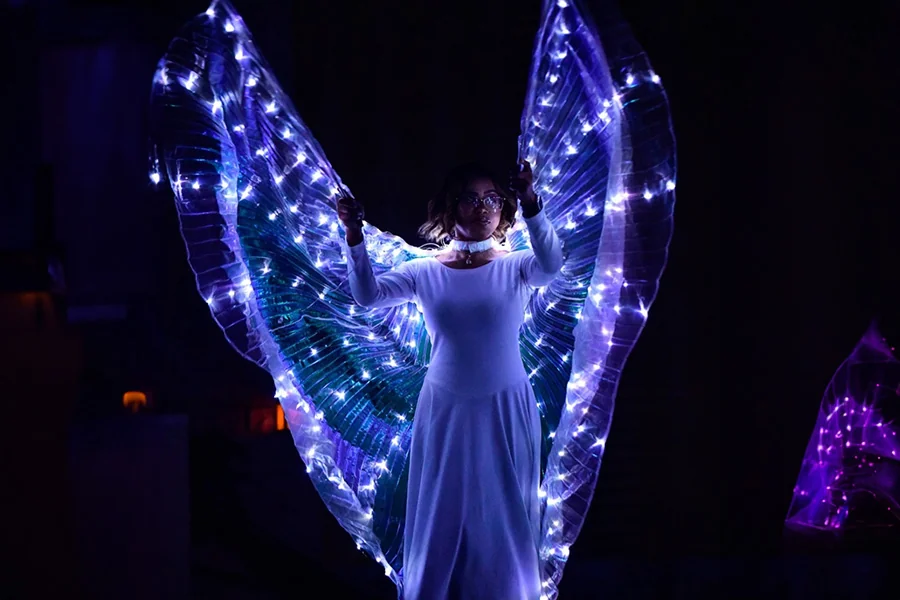 A performer with illuminated wings spread across the dark stage